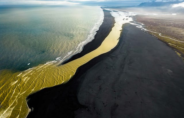 Glacier river with yellow color fall into the ocean coast in Iceland. Aerial view flying above yellow glacier river falling into the ocean, the yellow color in contrast with the volcanic landscape, the black sand beach and the horizon view. Iceland.